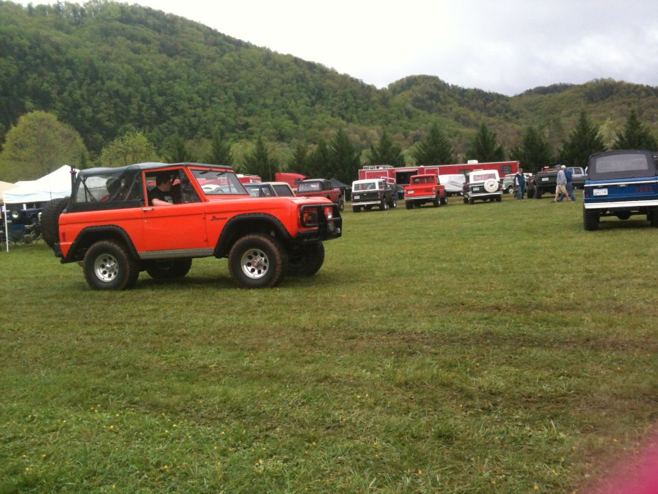 Orange first-gen Ford Bronco parked in a grassy field with other vehicles and tents in the background