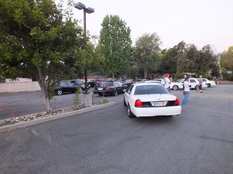 2003 Ford Crown Victoria - White full-size Ford sedan maneuvers in a parking lot as people watch at an evening gathering