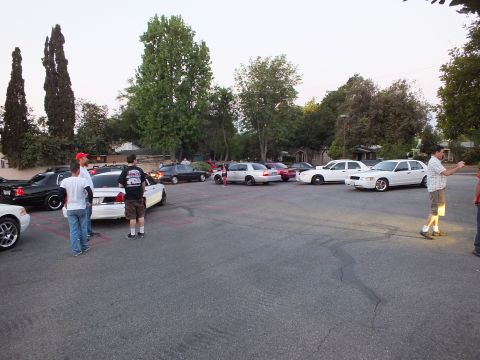 2003 Ford Crown Victoria - Group of people stand around an assortment of parked sedans across a large lot at dusk