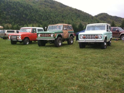 1992 Ford Bronco - Three classic Ford Broncos are parked on a grassy field with hills in the background.
