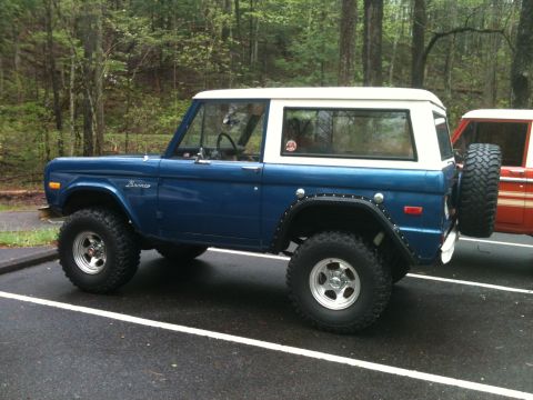 1992 Ford Bronco - Side view of a lifted blue Ford Bronco parked in a lot with trees in the background.