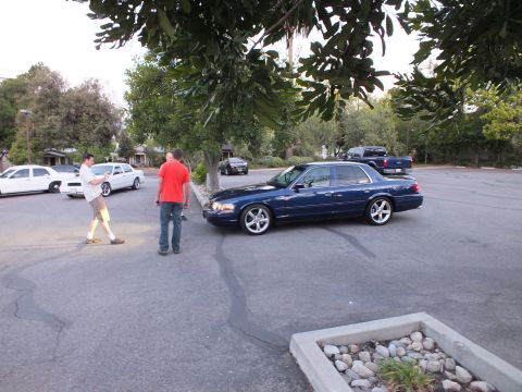 2003 Ford Crown Victoria - Dark blue full-size Ford sedan with chrome wheels rolls through a parking lot as two men walk past