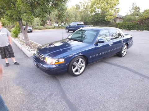 2003 Ford Crown Victoria - Front quarter of a dark blue full-size Ford sedan with chrome wheels parked along a tree-lined street