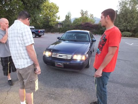 2003 Ford Crown Victoria - Men gather around a dark blue Ford Crown Victoria sedan at dusk in a lot, lights on