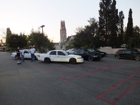 2003 Ford Crown Victoria - Several Crown Victoria and hatchback cars line a parking lot beneath a church bell tower