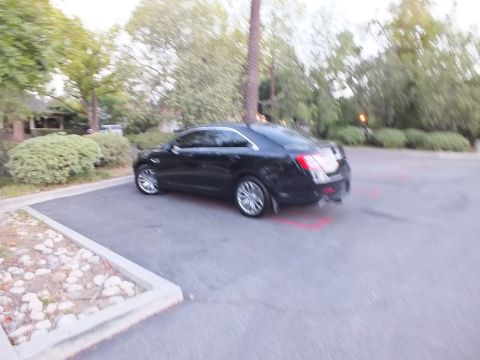2003 Ford Crown Victoria - Blurred black sedan rounds a red-striped parking lot drive lined with shrubs and rocks