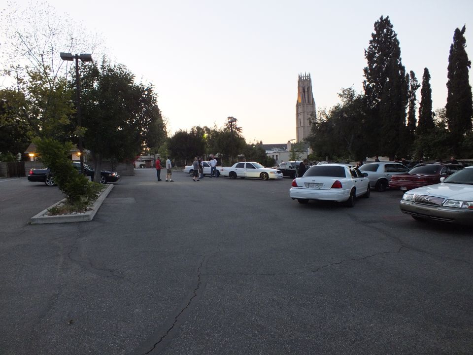 People stand near parked sedans in a large lot beside a lit church bell tower at dusk