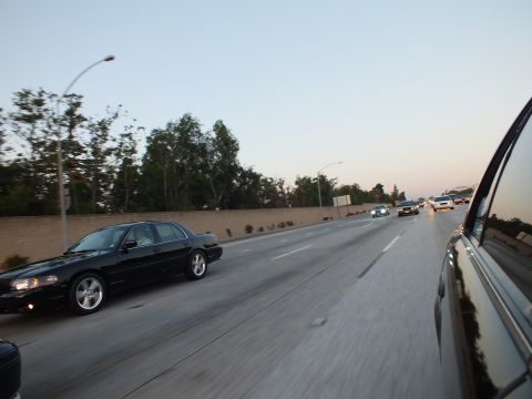 2003 Ford Crown Victoria - Black sedan rolls along an open freeway lane at dusk, shot from a passing vehicle