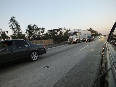 2003 Ford Crown Victoria - Gray sedan cruises a freeway beside a semi pulling a white RV trailer at dusk