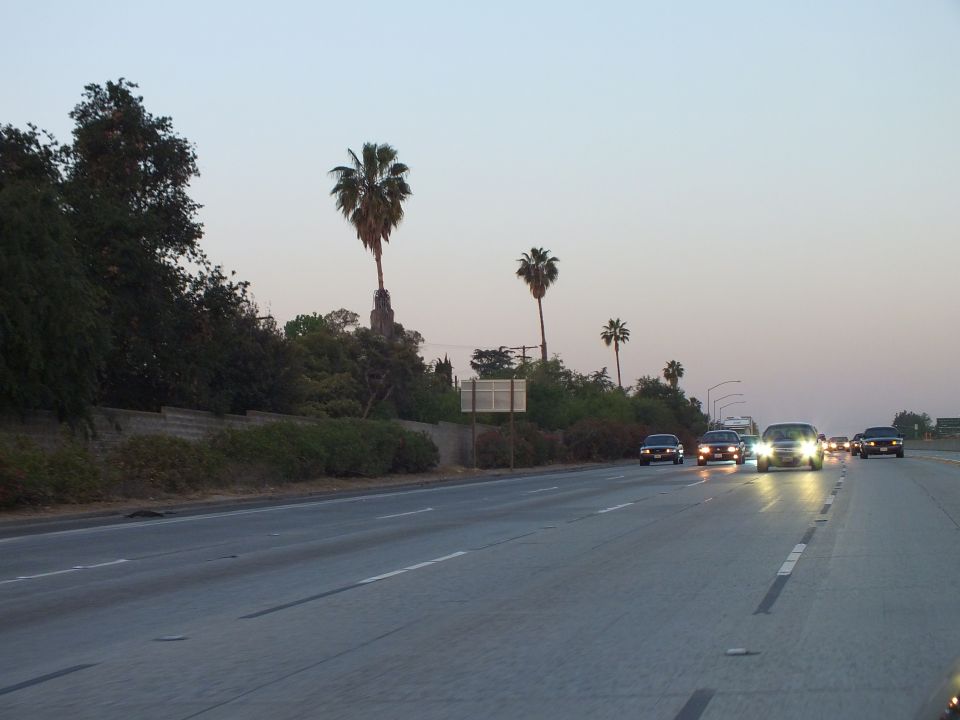 Freeway lanes at dusk with cars and headlights passing palm trees and a sign
