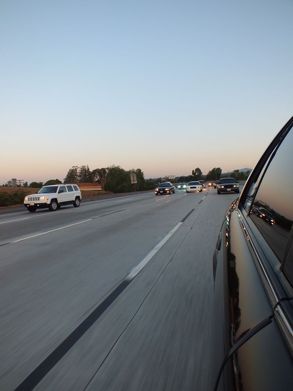 White Jeep Patriot SUV in left lane of freeway at dusk, dark car reflected at right