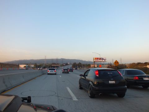 2003 Ford Crown Victoria - Ford Focus and full-size sedan move through right freeway lanes beneath a billboard.