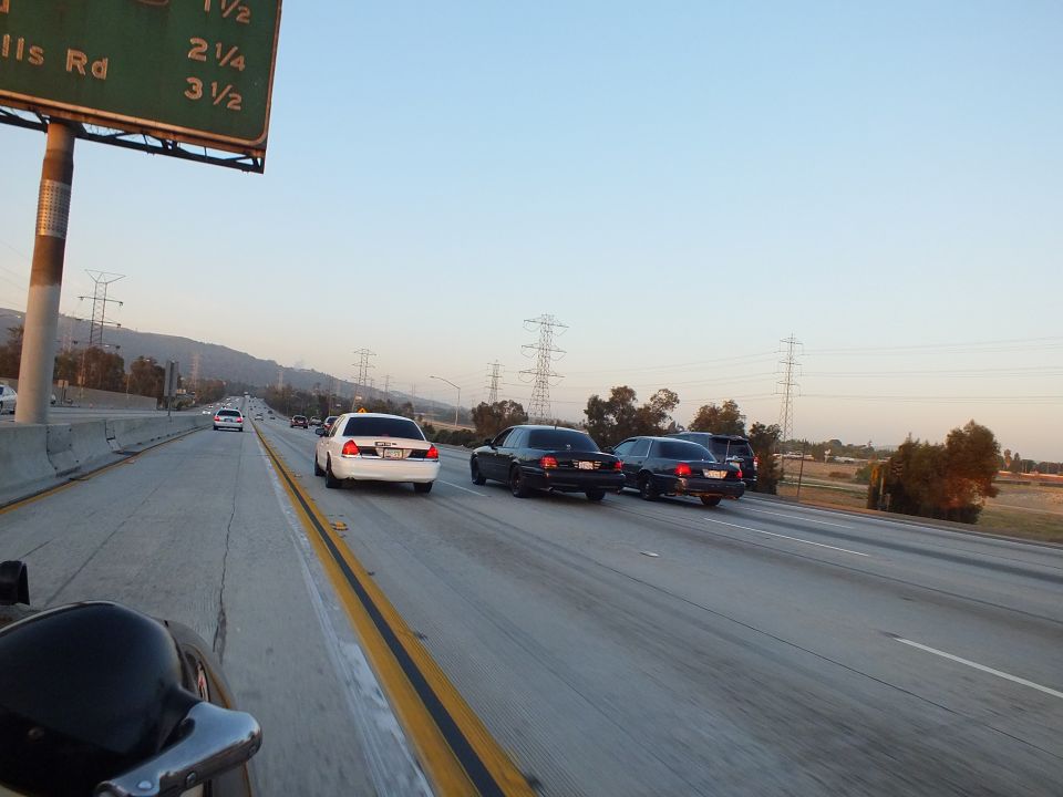 White and black full-size sedans grouped on a freeway below a green exit sign