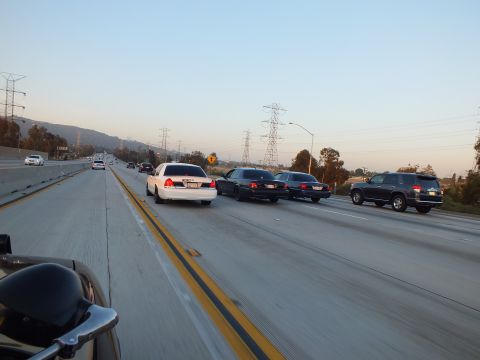 2003 Ford Crown Victoria - white full-size sedan and dark SUV cruise the freeway past hills and power towers.