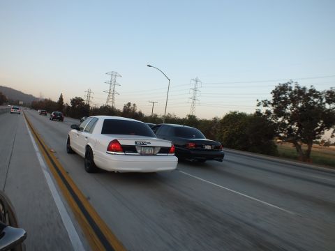 2003 Ford Crown Victoria - Two sedans run side by side down the freeway past steel transmission towers at dusk.