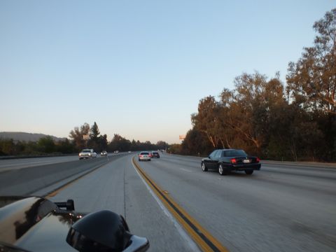2003 Ford Crown Victoria - black full-size sedan travels the right freeway lane beside a wall of trees.
