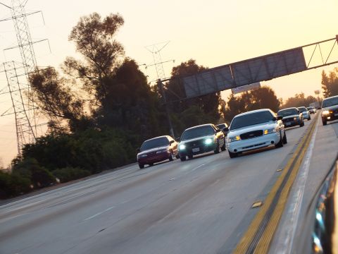 2003 Ford Crown Victoria - pack of cars rolls under a sign gantry with headlights lit against the setting sun.
