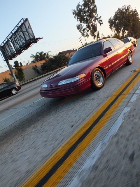 2003 Ford Crown Victoria - maroon sedan rolls past an empty billboard frame, framed by a double yellow line.