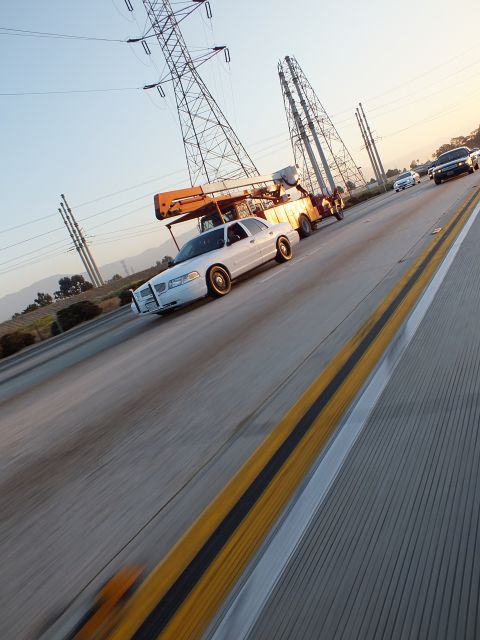 2003 Ford Crown Victoria - Low-angle motion shot of a white car overtaking a bucket-truck on the highway shoulder.