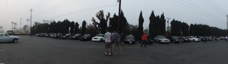Wide panorama of a large parking lot full of cars with people gathered under gray sky