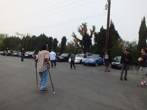 2003 Ford Crown Victoria - man on crutches stands in the lot as attendees gather near parked cars and a coupe.