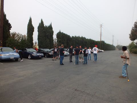 2003 Ford Crown Victoria - Attendees cluster on the asphalt near a row of dark sedans, with one man holding a tool at right.