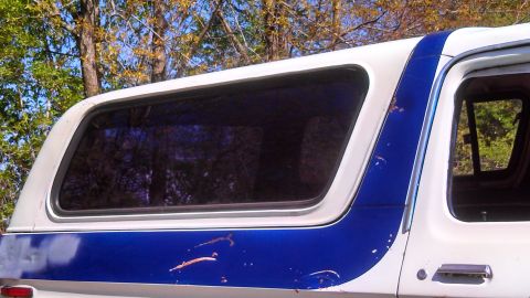 1979 Ford Bronco - The side of a pickup truck with a camper shell is shown, a blue stripe and white paint.