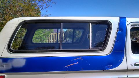 1979 Ford Bronco - The side of a white pickup truck with a blue stripe and a camper shell is shown under a clear blue sky.