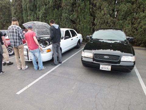 2003 Ford Crown Victoria - Plate 6NYR351 on a black Crown Victoria; a white example with raised hood draws a small crowd.