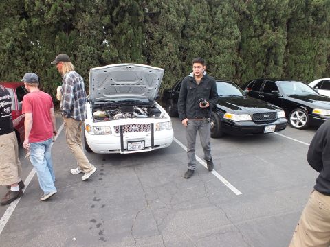 2003 Ford Crown Victoria - Man stands in the lot beside a white Crown Victoria with open hood, black sedans parked at right.