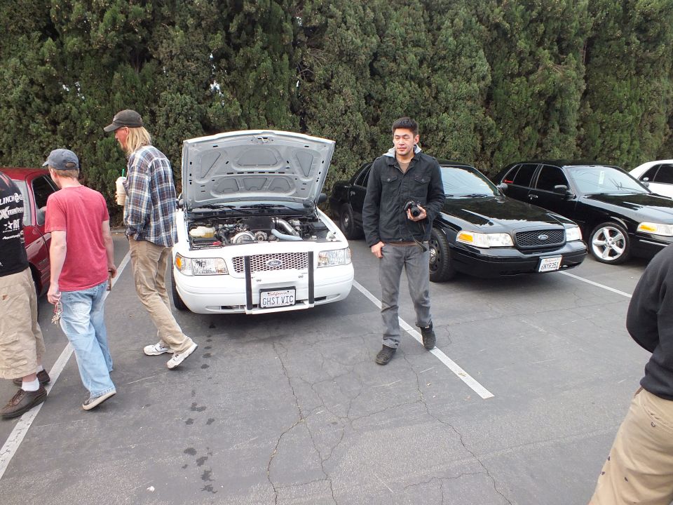 White Ford Crown Victoria with hood raised among parked sedans and people