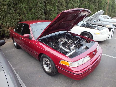 2003 Ford Crown Victoria - Dark red Crown Victoria on chrome wheels displays its turbo engine, white car parked behind.