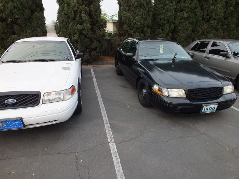 2003 Ford Crown Victoria - Two Ford Crown Victoria sedans, one white with Arizona plate and one black with Nevada plate, in adjacent parking spaces