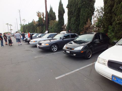 2003 Ford Crown Victoria - Lineup of Ford sedans including Crown Victoria and a black Focus along a parking lot row with spectators