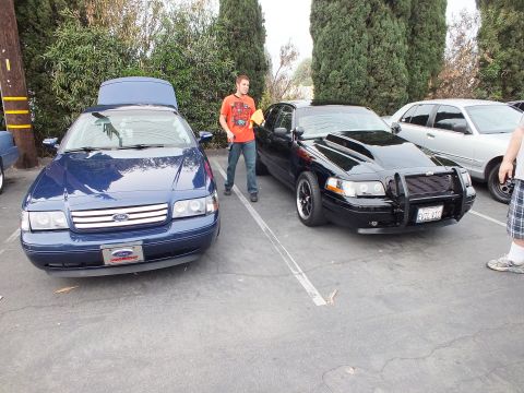 2003 Ford Crown Victoria - Dark blue Ford Crown Victoria with hood up next to a black push-bar Crown Victoria, man in orange shirt between them