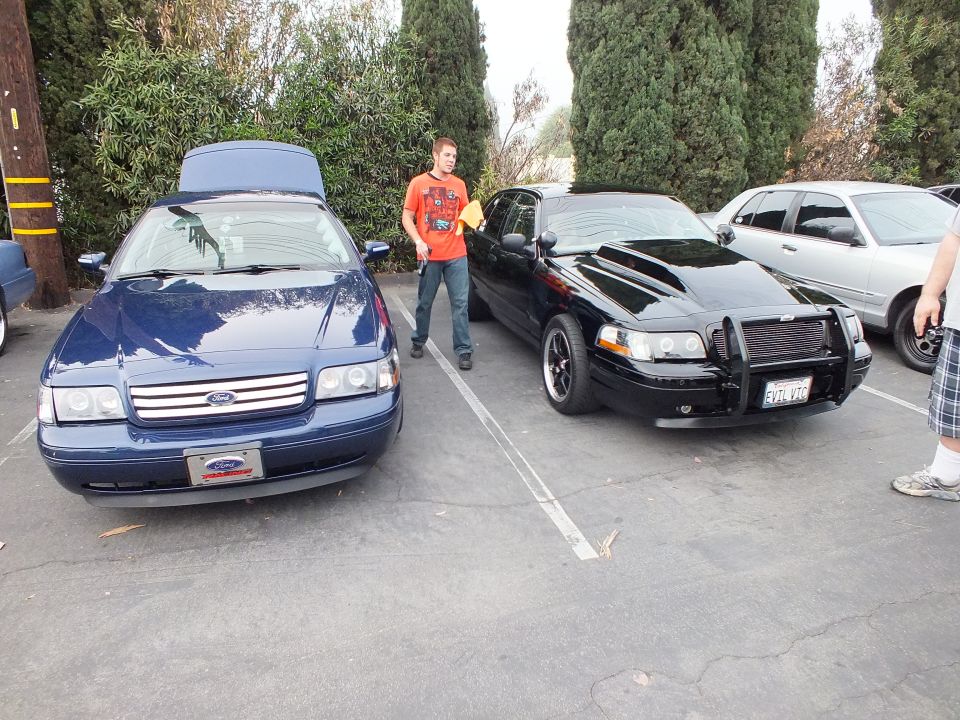 Blue Crown Victoria with open hood beside black push-bar Crown Victoria, man in orange shirt