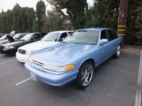 2003 Ford Crown Victoria - Light blue first-gen-style Ford Crown Victoria with large chrome wheels parked beside white and gray sedans