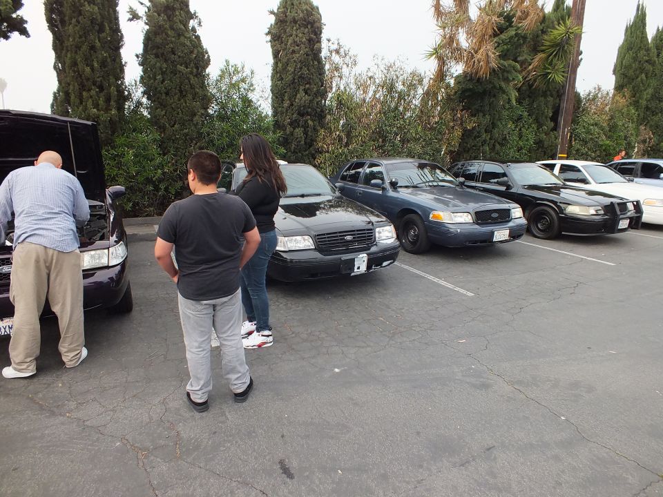 Dark Ford Crown Victoria sedans parked with people inspecting an open-hood car