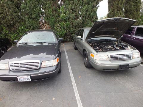 2003 Ford Crown Victoria - Gray Crown Victoria beside a green Crown Victoria with raised hood exposing the engine.
