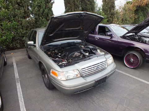 2003 Ford Crown Victoria - Sage-green Ford Crown Victoria displays its engine bay next to a purple sedan on wire wheels.