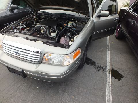 2003 Ford Crown Victoria - Front quarter of a silver Ford Crown Victoria, hood up over a modified intake, oil spot on pavement.