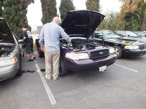 2003 Ford Crown Victoria - Lowered purple Ford Crown Victoria with hood up while attendees inspect nearby engine bays.