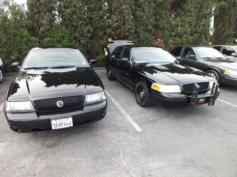 2003 Ford Crown Victoria - Black Mercury sedan parked next to a black Ford Crown Victoria equipped with a front push bar.