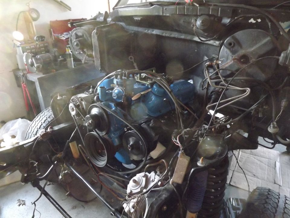 Engine bay with blue engine block, alternator, belts, and radiator. Wires and hoses are visible. Vehicle is in a workshop