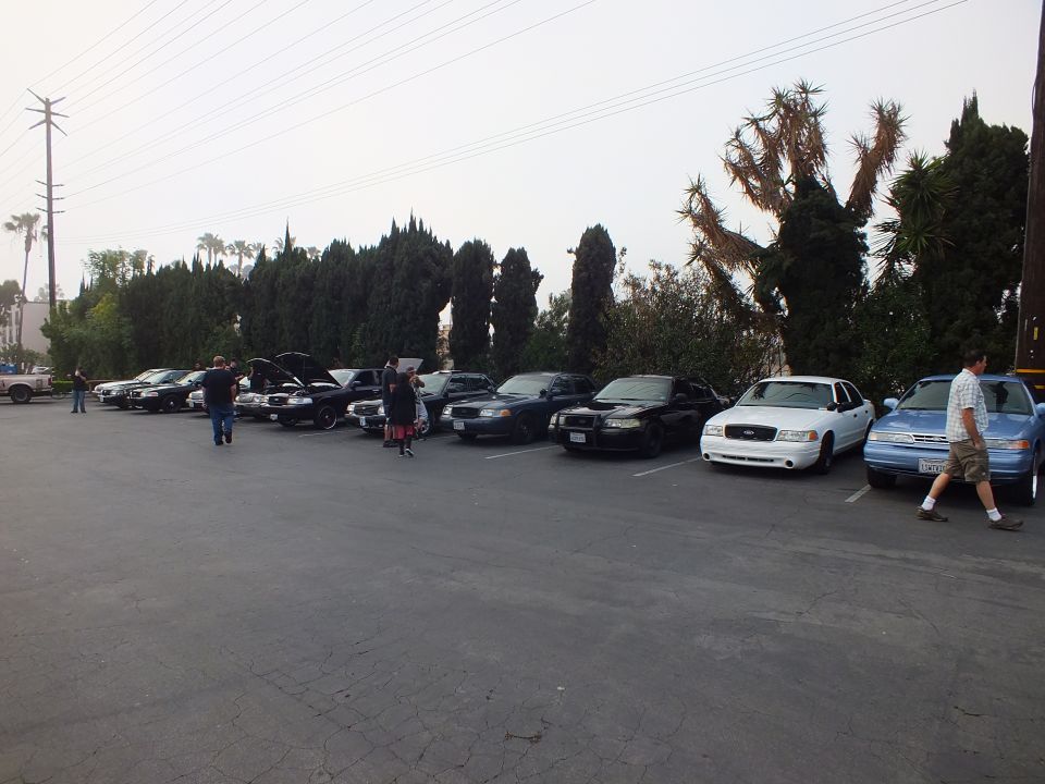 Row of sedans including white and blue cars parked at angle with people walking