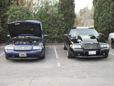 2003 Ford Crown Victoria - blue Ford sedan with its hood raised parked beside a black sedan fitted with a brush guard and an EVIL VIC license plate.