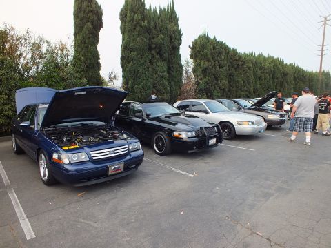 2003 Ford Crown Victoria - dark-blue Ford sedan with its hood raised parked beside black and silver full-size sedans, with people standing at right.