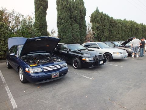 2003 Ford Crown Victoria - blue Ford sedan with raised hood parked next to a black sedan and a silver sedan as people stand at the right.