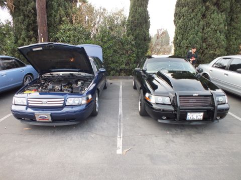 2003 Ford Crown Victoria - blue Ford sedan with hood raised parked beside a black sedan with a brush guard and an EVIL VIC license plate.