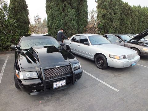 2003 Ford Crown Victoria - black sedan fitted with a brush guard and EVIL VIC plate parked next to a silver Mercury Grand Marquis at a car meet.